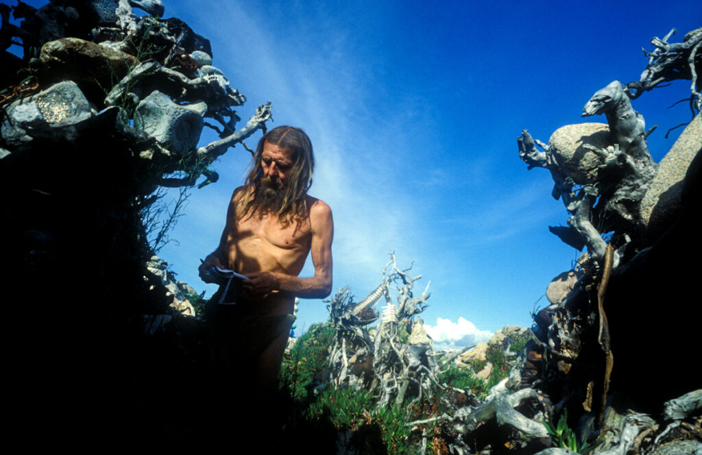 Manfred Gnädinger standing shirtless among organic stone sculptures in Camelle’s coastal landscape.