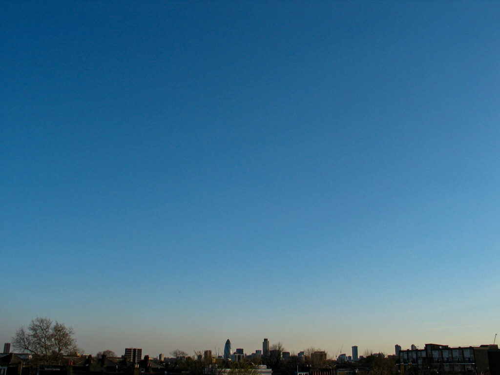 Clear blue sky stretching over the London cityscape at sunrise or sunset, with buildings and trees silhouetted along the horizon and minimal clouds present.