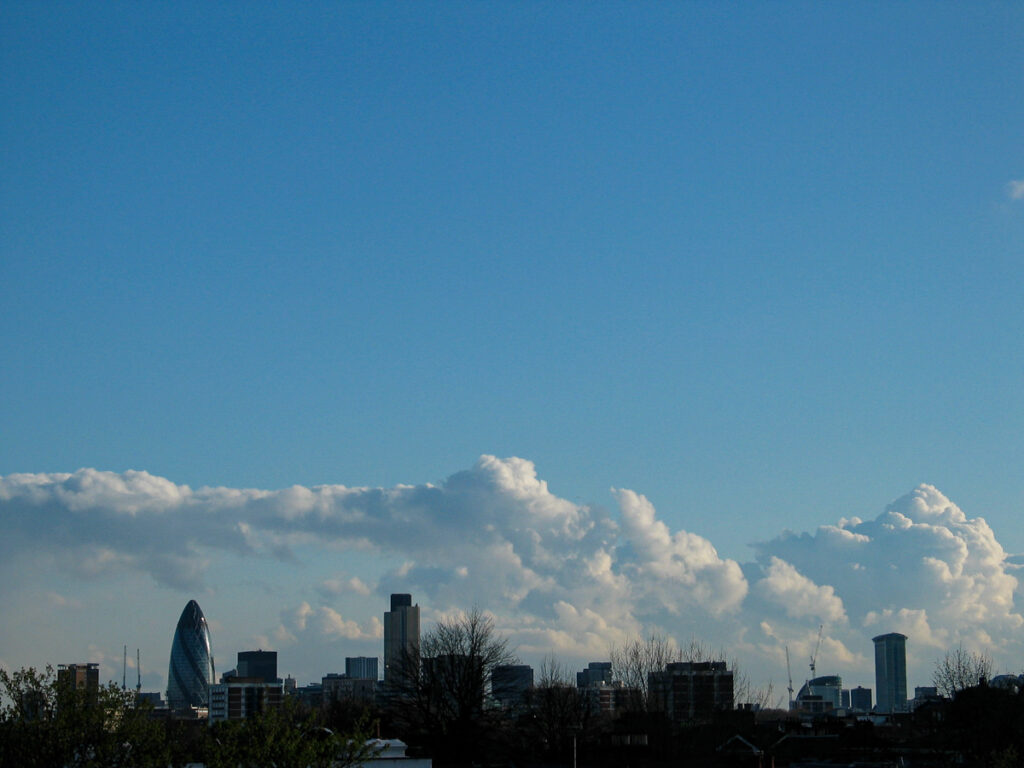 City of London skyline seen from Hackney during the evening on Monday 19 April 2004, with The Gherkin, Tower 42, and One Canada Square silhouetted against a blue sky featuring cumulus and stratocumulus clouds.