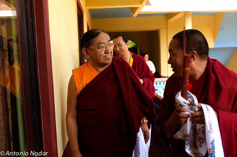 Monks in maroon and saffron robes stand near a sunlit hallway, holding ritual objects wrapped in white ceremonial scarves.