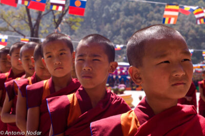 A line of young monks in red and saffron robes stand outdoors beneath rows of colorful flags during a monastery ceremony.