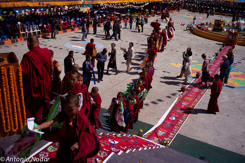 Monks and novices in colorful robes walk in line along decorated carpets and steps during a monastery opening celebration, with crowds gathering in the background.