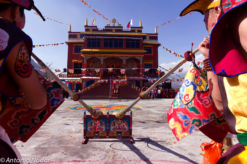 Two ritualists in ornate costumes stand before giant Tibetan horns, aimed toward the grand, colorful monastery facade during a celebration.