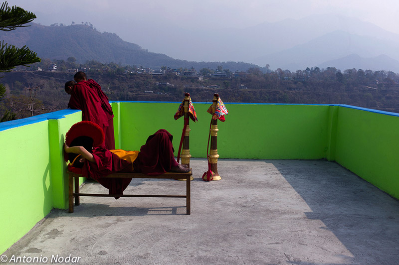 Two monks in maroon robes, one reclining on a bench and the other leaning on a bright green wall, overlook a misty valley with ceremonial instruments nearby.