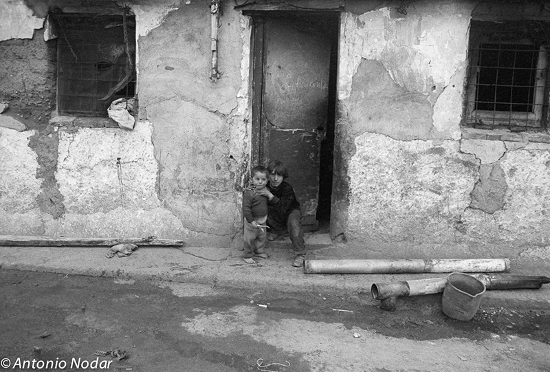 Two children sit and stand outside a crumbling entrance to a humble home, with exposed pipes and a bucket on the ground in Perona, Barcelona.