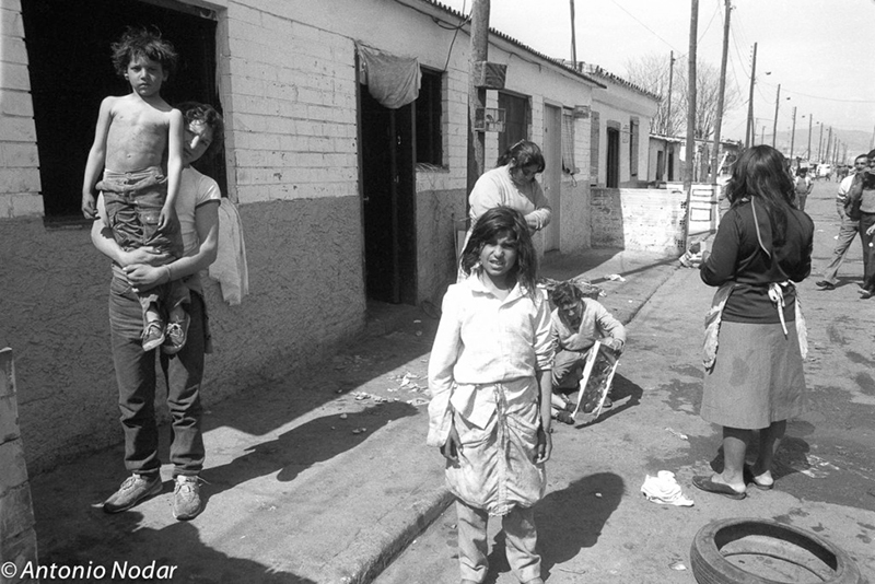Adults and children gather along a street outside modest homes, with one child riding on an adult's shoulders and another standing in the foreground, illustrating daily life in Barcelona's Perona shanty town.