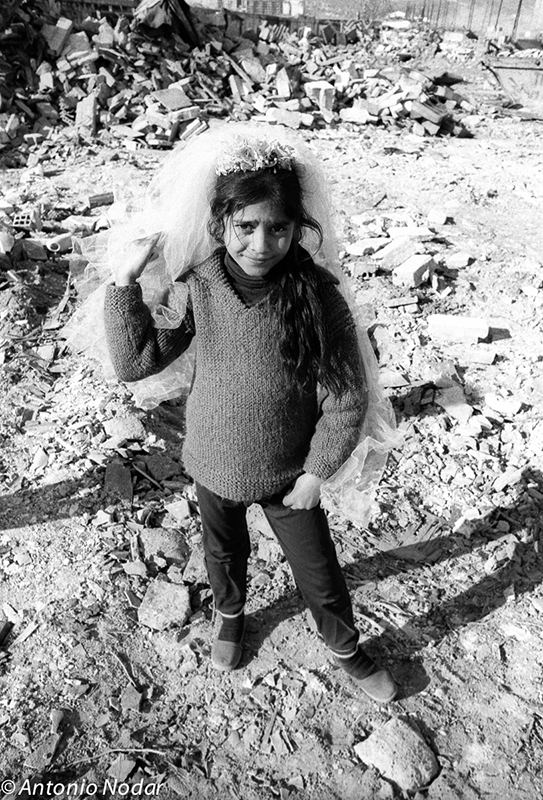 A young girl stands in a debris-strewn lot, wearing a bridal veil and sweater, gazing directly at the camera in the Perona shanty town of Barcelona.