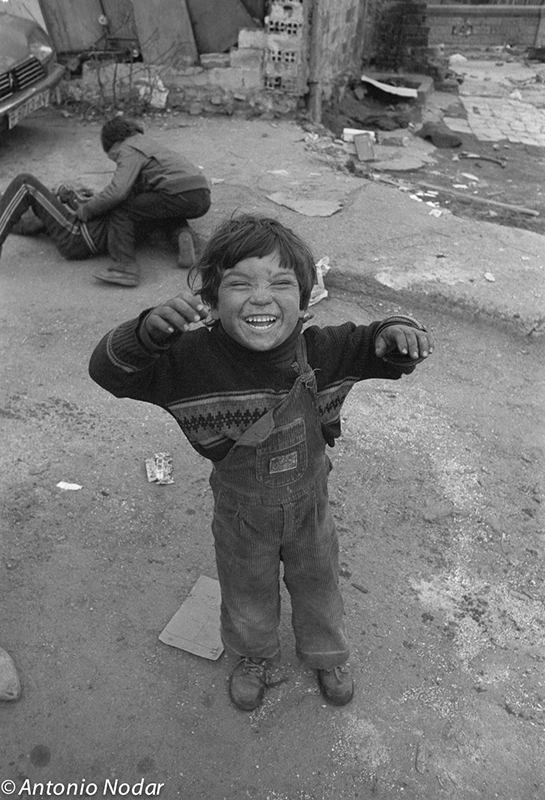 A smiling child in overalls stands on a rough street, arms raised playfully, with another child in the background amid the debris of La Perona, Barcelona.