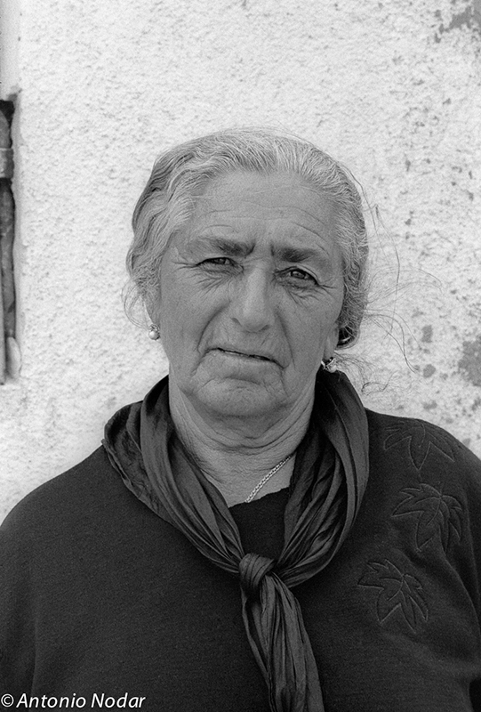 Close-up portrait of an elderly woman with white hair and a scarf, standing against a textured wall in Barcelona’s Perona neighbourhood.