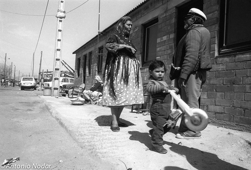 A child plays with a toy scooter on the sidewalk while an older woman and man stand conversing, with more residents visible outside brick homes in Barcelona’s Perona neighbourhood.