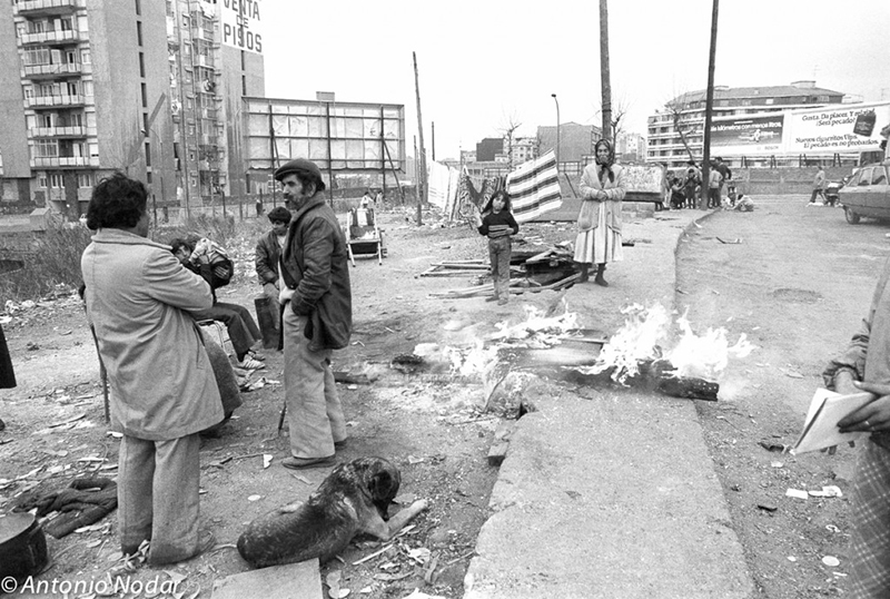 Residents interact around a makeshift street fire, with a dog lying nearby and scattered debris, set against high-rise buildings bordering the Perona former shanty town in Barcelona.