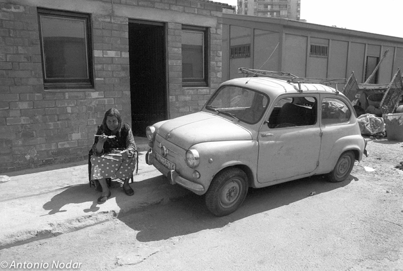 A woman sits on a roadside in front of a small vintage car parked outside simple brick dwellings, with urban buildings visible in the distant background.