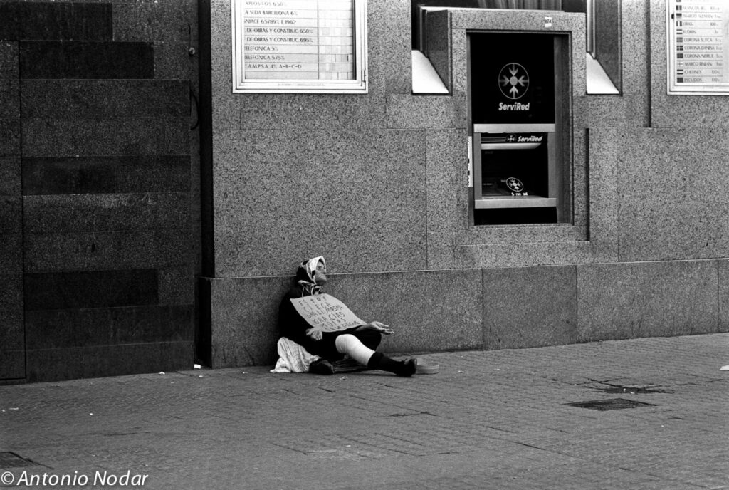 Elderly woman sits on the pavement by a city bank, holding a handwritten sign beside an ATM, Barcelona, 1990s.
