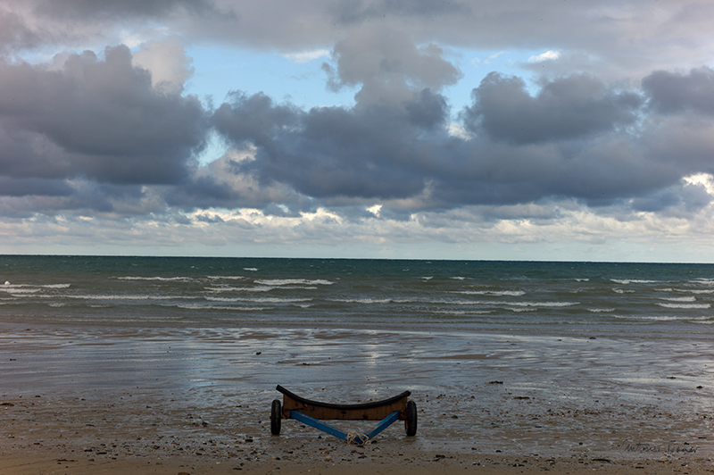 An empty boat trolley stands on the wet sand at low tide facing the sea under dramatic clouds in Coutainville, Normandy.