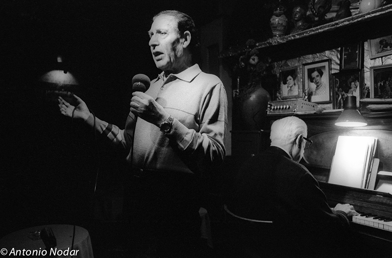 A singer performs beside a pianist in a dimly lit bar, surrounded by photographs and keepsakes.
