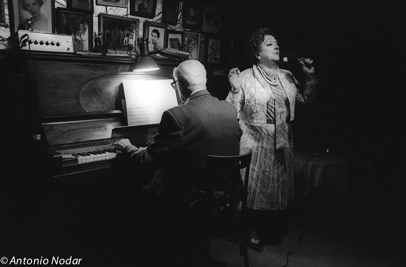 A woman in elegant attire sings into a microphone while a pianist plays under a glowing lamp, surrounded by framed photos.