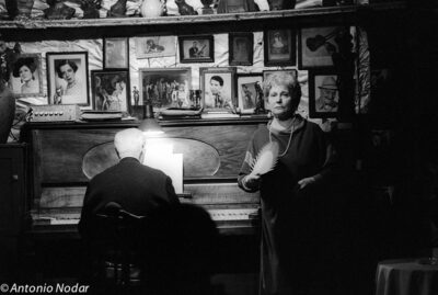 A woman holding a fan stands beside a pianist at an upright piano, surrounded by framed photographs.