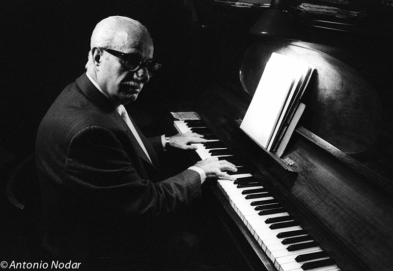 An elderly pianist in a suit sits at an upright piano, hands hovering over the keys under a soft lamp.