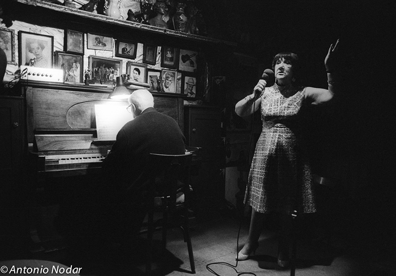 A performer stands beside a pianist, microphone in hand, in front of a wall densely decorated with photographs.