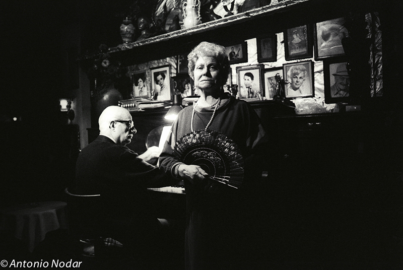 A woman with a decorative fan stands beside a pianist, illuminated by soft light and surrounded by framed photos.
