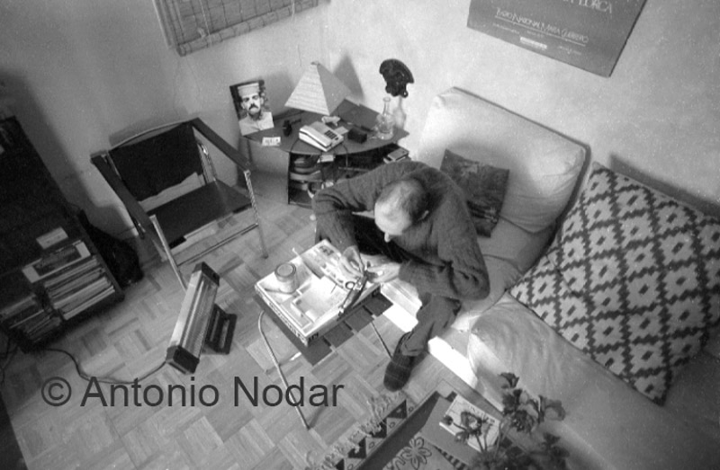 Carlos Moro sits alone in his living room, surrounded by personal belongings, framed by patterned pillows and posters.