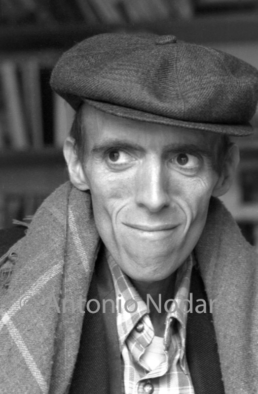 Carlos Moro wearing a flat cap and scarf, sitting indoors in Barcelona, facing the camera in a moment of calm.