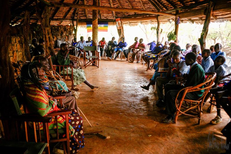 Community members of all ages gather in a rustic, open hall for a group meeting, seated in a circle of chairs, with vibrant village attire and lively interaction.