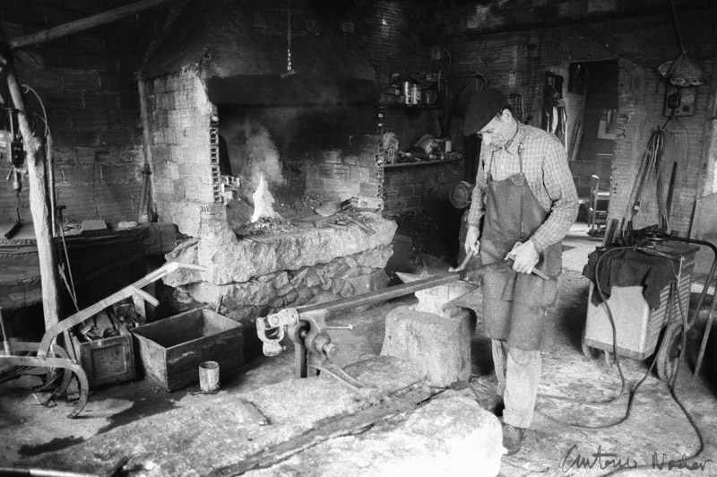 A blacksmith in an apron and cap works with metal tools beside a glowing forge inside a rustic, stone-walled workshop.