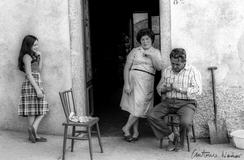 A girl in a plaid dress leans against a wall while a woman and a man sit and stand at the entrance of a rural home, with a shovel and bowl nearby.