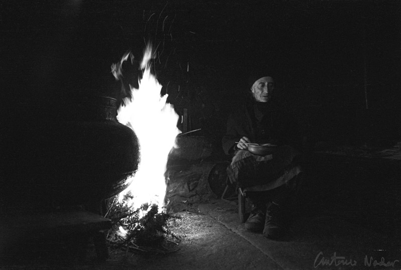 An elderly woman wearing a patched and well-worn work blouse and headscarf stands in deep shadow, illuminated by a beam of light from behind.
