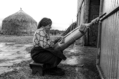 An elderly woman sits on a small stool in a farmyard, skillfully working at a traditional bobbin lace pillow, with large haystacks visible in the background.
