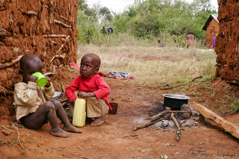 Two small children sit at the base of a weathered clay wall, drinking and eating. A kettle simmers on makeshift firewood beside them, filling the air with smoke and promise of food.