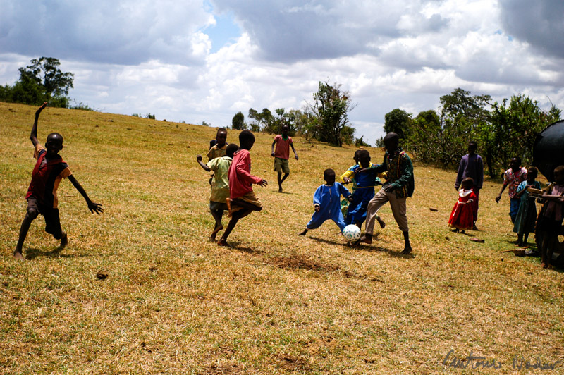 Children chase after a soccer ball in a golden field while others watch or cheer nearby. The scene is lively, full of movement and laughter, under a sky filled with dramatic clouds.