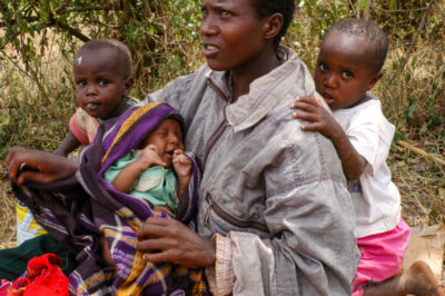 An adult wearing a gray jacket holds a bundled infant while surrounded by several young children outdoors, beneath dense foliage. The children gather closely, some leaning on the adult for support, while others reach out with gentle hands.