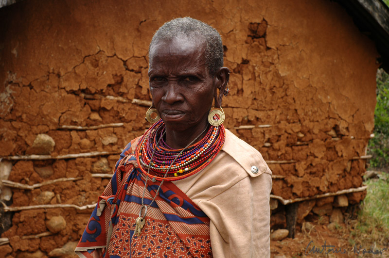 A woman wearing layered, colorful beaded necklaces and patterned fabric stands before a rustic mud-walled house, adorned in traditional attire and jewelry.