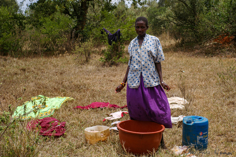 A woman stands in a dry grassy field, surrounded by scattered clothing, buckets, and a blue water container, engaged in daily chores beneath a canopy of distant trees.