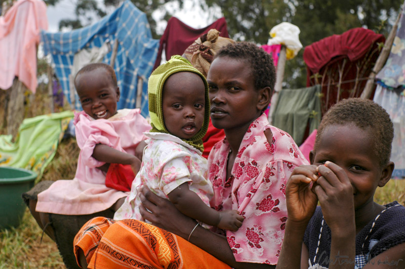 A woman dressed in a vibrant patterned wrap sits on the ground holding an infant, while two small children—one in a pink dress—sit nearby. Behind them, colorful clothes hang on a makeshift line in an open, grassy area.