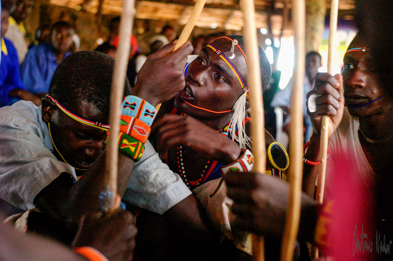 A group of people in traditional beadwork bracelets and necklaces grip ceremonial sticks together inside a meeting house, deeply engaged in a communal ritual or discussion.