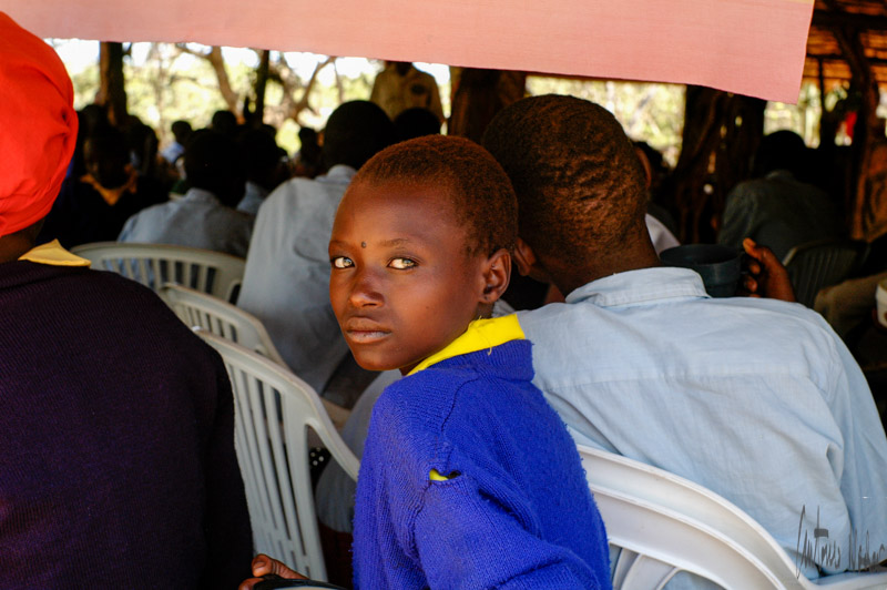 A child in a bright blue sweater and yellow collar sits beside an adult, surrounded by fellow students on white plastic chairs inside a shaded open-air classroom.