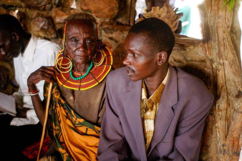 An elder adorned in multi-layered, colorful beadwork necklaces sits beside a younger man in a suit jacket inside a rustic, timber-and-stone hut, symbolizing the meeting of tradition and contemporary life.