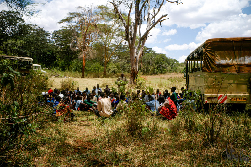A group of people from different age groups gather outdoors in a circle under tall trees beside two green trucks, engaged in a communal meeting in a grassy clearing.