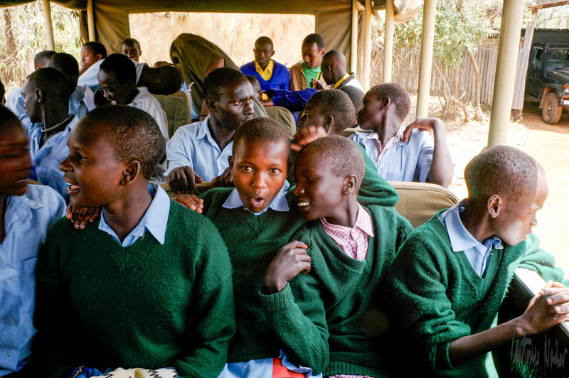 Students from local Kenyan tribes, dressed in green sweaters and blue shirts, sit together on benches inside an open-sided vehicle, prepared for a learning journey.