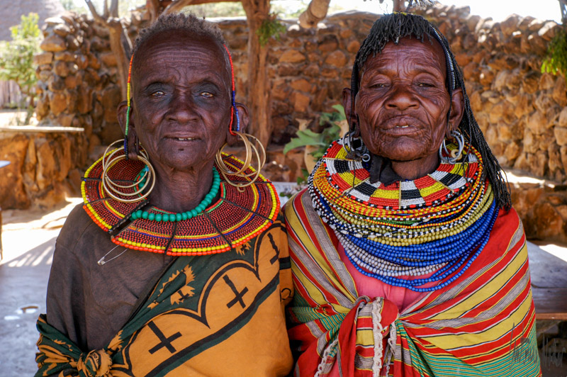 Two women from local Kenyan tribes stand together outdoors, adorned in vibrant, layered beadwork collars and patterned shawls, exemplifying traditional dress.