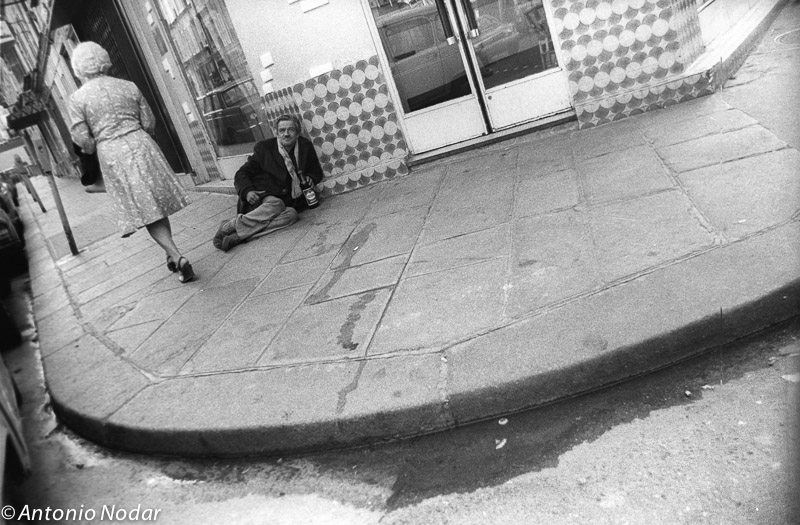 An elderly woman walks past a man sitting on a city sidewalk in Paris in the 1980s, while he holds a drink and looks away.
