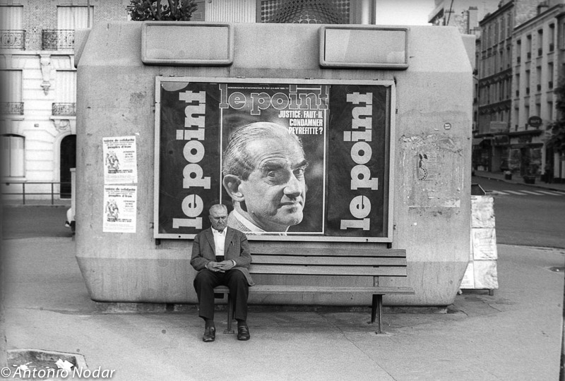 An elderly man sits alone on a bench in front of a large advertisement-covered structure in Paris, 1980s.