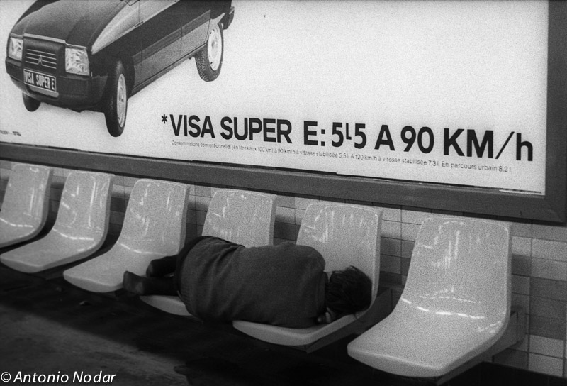 An older person lies curled on a row of plastic seats in a Paris metro station below a large advertisement for a car, 1980s.