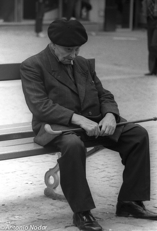 An elderly man in a beret sits on a park bench in Paris, 1980s, holding a cane and looking down.