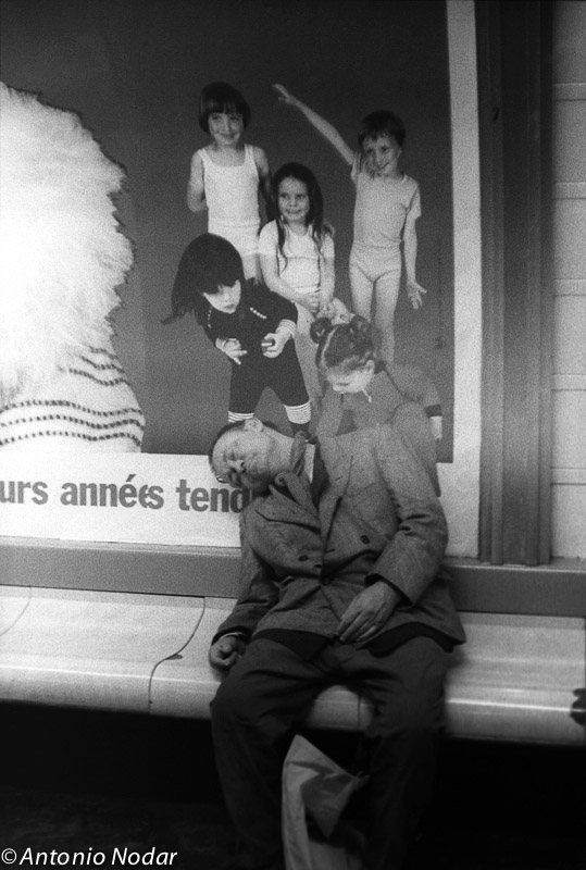 An elderly man sleeps slouched on a metro bench in Paris, 1980s, under a large advertisement featuring children.