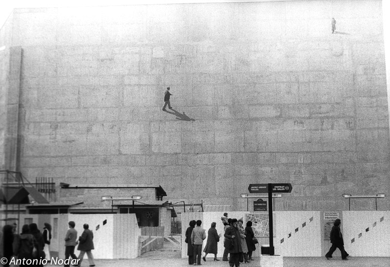 People walk past a large blank wall in Paris, 1980s; two small figures and a shadow are painted high on the wall.