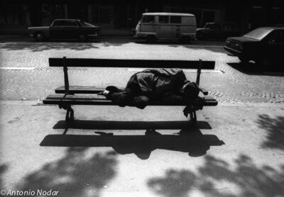 An elderly figure sleeps curled on a park bench in Paris, 1980s, surrounded by deep shadow and midday street light.